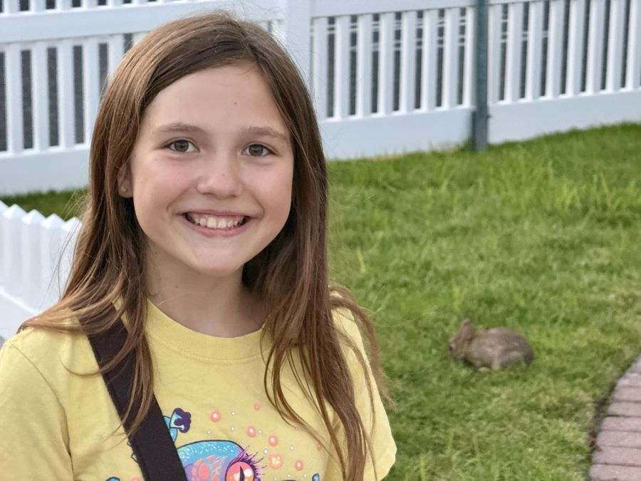 Emma smiling in a yellow shirt with a little bunny in the grass behind her